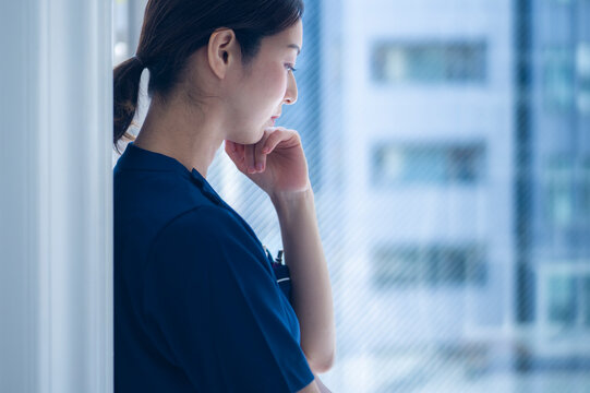 A female nurse in scrubs stands beside a window, her hand resting thoughtfully on her chin as she looks down, appearing deep in thought or concern.