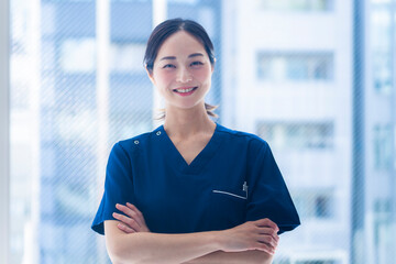 A young female nurse in blue scrubs stands confidently with folded arms inside a modern hospital...