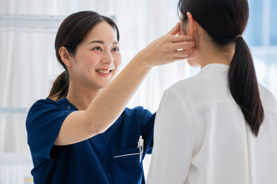 A female doctor in blue scrubs examines a female patient's facial features during a consultation in a bright medical clinic. The patient is holding still as the doctor uses a medical tool for assessme