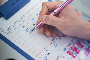 A close-up image of a doctor&rsquo;s hand writing patient notes or appointment details on a clinical chart with a pink pen during a consultation.