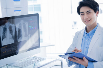 A male doctor wearing a white lab coat stands in a modern clinic, smiling while holding a...