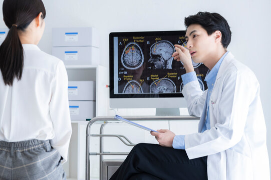 A male doctor in a white coat explains brain MRI images to a female patient during a neurological consultation in a bright clinic.

