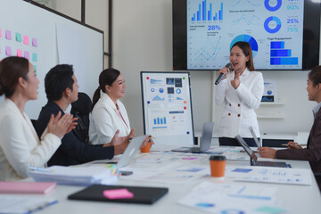Asian businesswoman leading a meeting, presenting with a microphone while colleagues listen attentively and applaud, as charts and graphs display on screens and flip charts