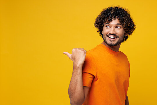 Side view young smiling Indian man wear orange t-shirt casual clothes point thumb finger back aside on blank empty area mockup isolated on plain yellow background studio portrait. Lifestyle concept.