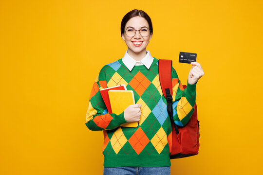 Young smiling happy smart girl student wears green casual clothes backpack bag hold books credit bank card isolated on plain yellow background studio portrait. High school university college concept.
