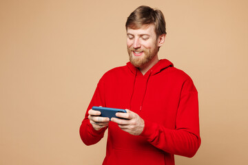 Young man he wear red hoodie casual clothes use play racing app on mobile cell phone hold gadget smartphone for pc video games isolated on pastel plain light beige background studio Lifestyle concept