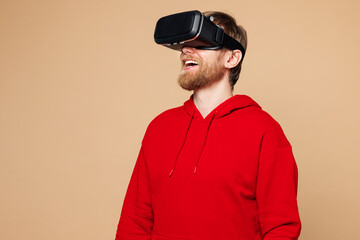 Young amazed Caucasian man he wearing red hoodie casual clothes watching in vr headset pc gadget look aside on area isolated on pastel plain light beige background studio portrait. Lifestyle concept.