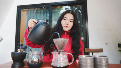 A woman in a red jacket pours hot water from a black kettle into a coffee dripper, preparing a fresh brew at a cozy indoor setting.