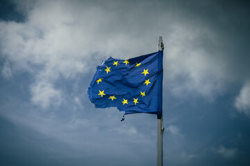 A torn EU flag blows in the wind with a few clouds in the background