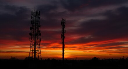 Naklejka premium Two communication towers stand silhouetted against a vibrant sunset with colorful clouds above them