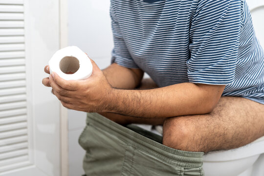 Male holding toilet paper roll and sitting on toilet in restroom. Man squeezing tissue with diarrhea excreting in toilet. Hand holding a roll toilet paper is feeling pain with constipation.