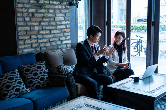 A businessman in a suit is holding a blue coffee cup and engaging in a lively discussion with a female colleague. They are seated in a cozy, modern caf&eacute; lounge with exposed brick walls, suggesting a c