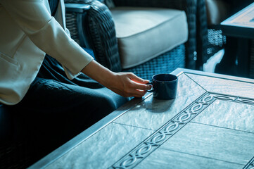 A close-up shot of a woman in a cream blazer reaching for a blue coffee cup on a tiled table in a stylish café. The focus is on her hand and the coffee cup, conveying a relaxed and quiet moment.