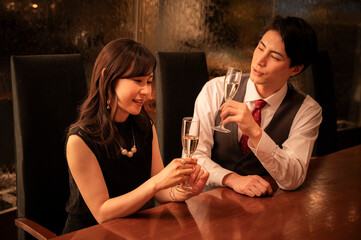 A man and woman dressed formally are smiling at each other while toasting with champagne glasses. They are seated across from each other at a wooden bar with warm ambient lighting.