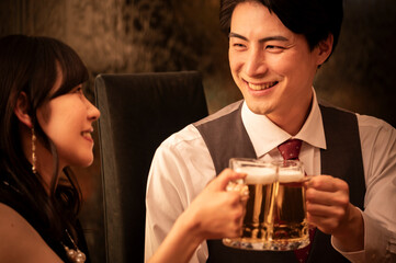 A cheerful couple in formal evening attire clinks beer mugs at a stylish bar. The man wears a vest and red tie, while the woman wears a sleeveless black dress. The scene is warmly lit and captures a j