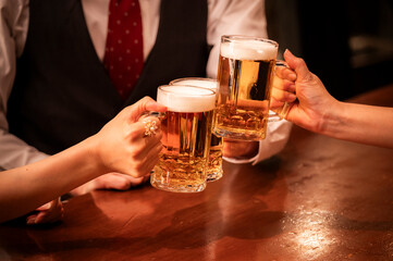A warm-toned image of 4 people clinking beer mugs at a modern bar. The close-up focuses on their hands and glasses filled with golden beer, capturing a festive group moment.