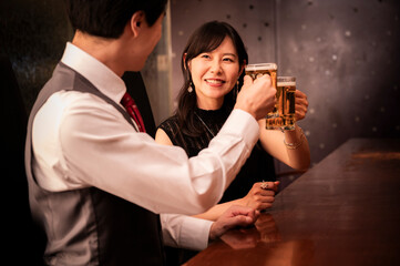 A cheerful couple in formal evening attire clinks beer mugs at a stylish bar. The man wears a vest and red tie, while the woman wears a sleeveless black dress. The scene is warmly lit and captures a j