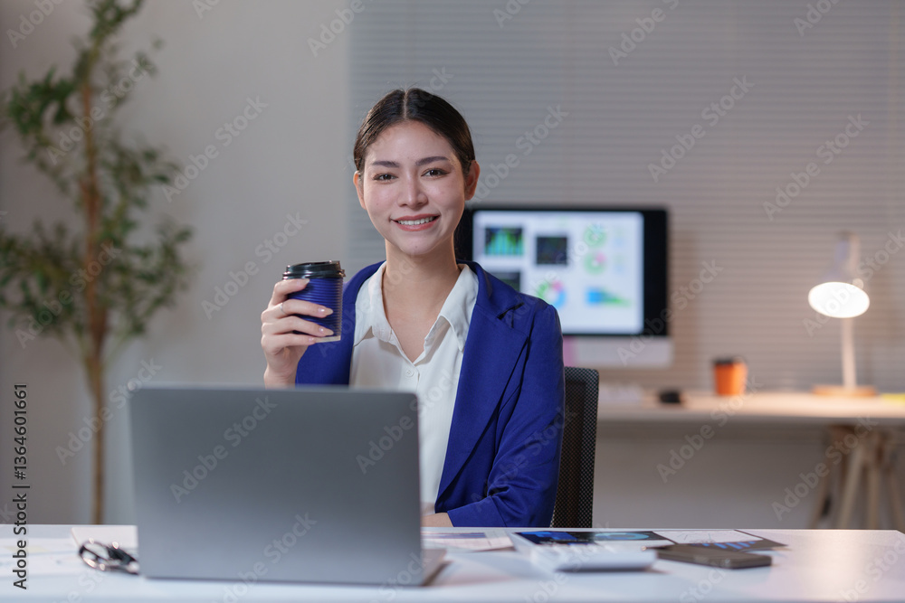 Poster smiling businesswoman drinking coffee while working late at night on a laptop in a modern office wit