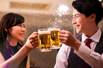 A joyful man and woman clink their beer mugs together in a celebratory toast at a warmly lit bar. Both are smiling brightly, creating a cheerful and relaxed atmosphere.