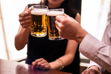 A close-up shot of a man and woman toasting with beer mugs in a warmly lit bar. The image focuses on their hands and glasses, emphasizing the golden color of the beer and the festive atmosphere.