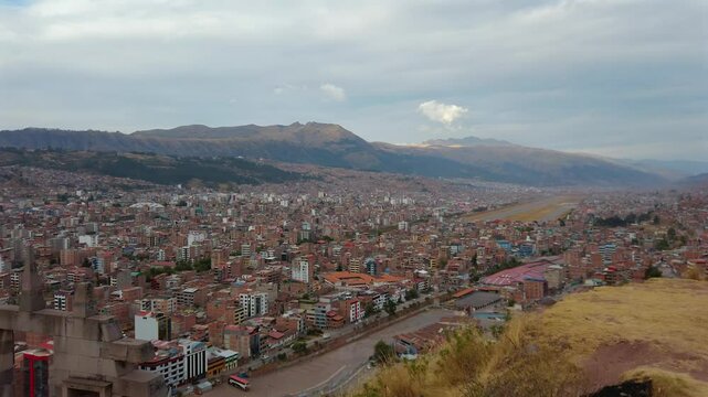 Pan right from Mirador Pachacutec reveals the Puerta del Sol monument, sprawling city of Cusco, and visible airport runway nestled in the Andean landscape under soft daylight.