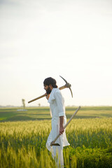A hardworking Pakistani Indian farmer walks through a lush green field, carrying a pickaxe and hoe on his shoulders at sunrise.