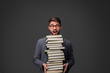 A surprised young man holds a large stack of books against a dark background. His expression...