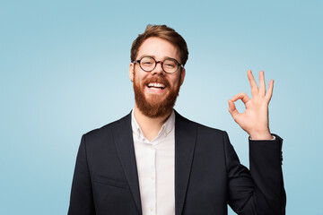 A happy professional man with glasses smiling warmly and making an OK hand sign, displaying confidence and positive energy. Captured against a soft blue background.