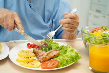 Asian senior woman patient eating pork chop stake and vegetable salad for healthy food in hospital.