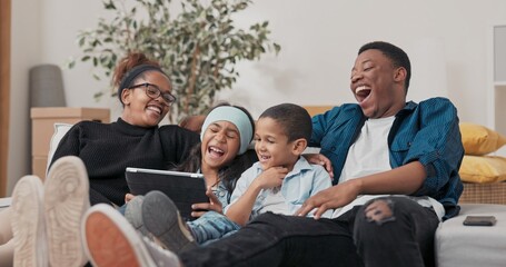 A joyful family sits on a sofa in their freshly bought house. The kids watch cartoons on a tablet while parents hug them and laugh together.