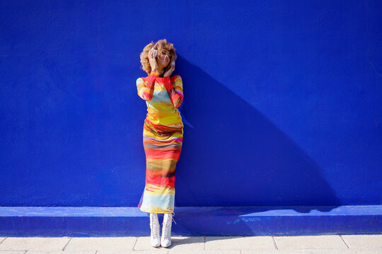 Woman with curly afro hairstyle dancing happily against a blue wall while listening to music on headphones