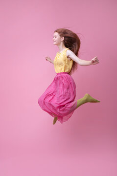 Red-haired girl jumping in a studio with a pink background wearing a long skirt and green tights