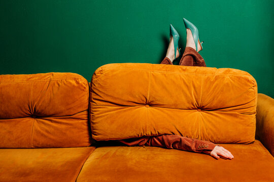 Woman in heels partially hidden in an orange sofa against a green wall, symbolizing tiredness and depression