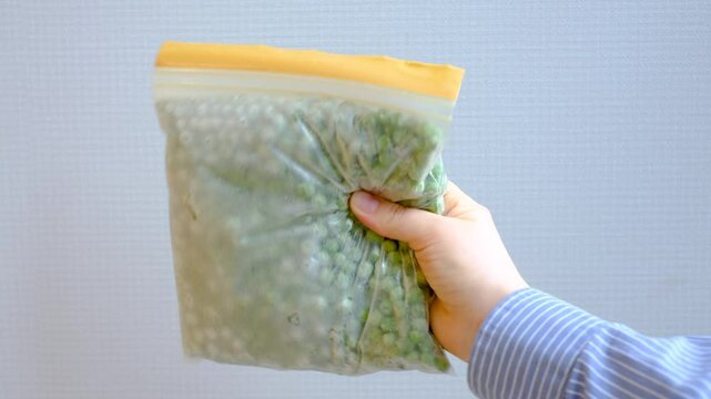 Female hand holding zip bag with frozen green peas on gray background close-up.
