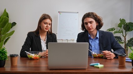 Business team of man and woman sitting at desk in the office having dinner break with food delivery, people talking working on laptop eating healthy salads.