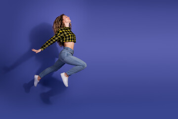 Stylish young woman jumping against purple background in casual checkered shirt and jeans