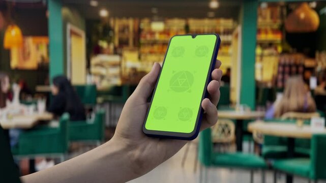 GREEN SCREEN CHROMA KEY Hand holding smartphone with green screen in a cozy cafe, ideal for app mockups and branding in a lively restaurant environment