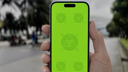 GREEN SCREEN CHROMA KEY Person holding a smartphone with a green screen outdoors near a tropical park and road, featuring palm trees and urban surroundings