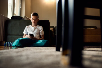 Boy sitting on floor at home using smart phone typing message to a friend.
