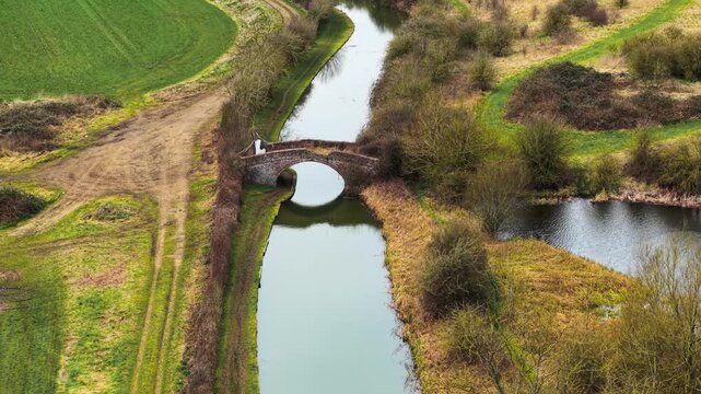 Historic brick bridge crossing narrow canal in United Kingdom. Calm water reflecting arched stone structure amid rural landscape. Old masonry spanning small navigation channel with grassy banks