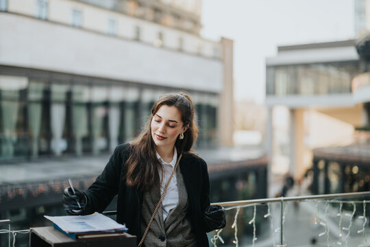 A young woman evaluates documents while standing on an illuminated balcony outdoors, with a modern cityscape in the background, signifying urban lifestyle, business professionalism, and focused work.