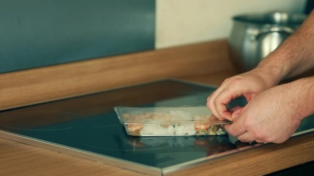 Opening plastic packaging of seafood mix on induction stove. Peeling off transparent film from container with assorted shellfish. Removing plastic cover from tray with shrimp, squid, and mussels