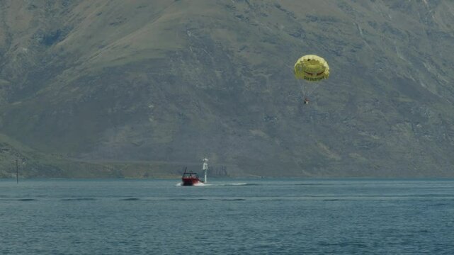 A parasailer is towed high above a lake by a red speedboat, set against steep, rugged mountains in Queenstown, New Zealand.