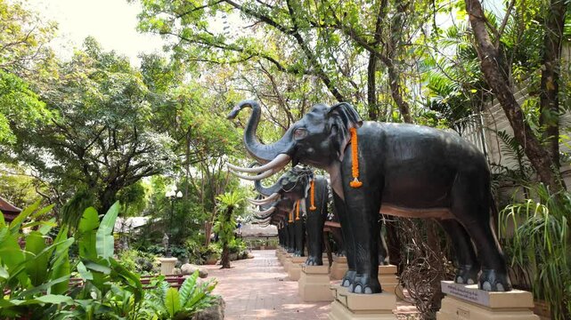 Forward-moving shot under a row of large black elephant statues adorned with orange garlands at Erawan Museum, Bangkok, with lush greenery and traditional architecture