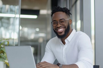 Obraz premium A man with glasses and a beard is sitting at a table with a laptop. He is smiling and he is happy
