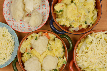 A top-down view of individual ramekins being prepared with mashed potatoes, cauliflower, leeks, and cheese, ready for baking into a gratin dish