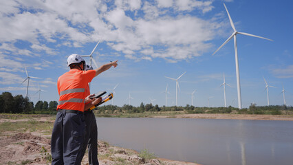 In a wind farm, two Asian inspectors are reviewing data on a tablet and clipboard to analyze system performance and blade rotation at varying wind speeds. Engineer Wind Turbine.