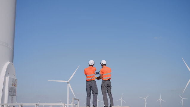In a wind farm, a rear view shows two Asian inspectors are reviewing data on a tablet and clipboard to analyze system performance and blade rotation at varying wind speeds. Engineer Wind Turbine.