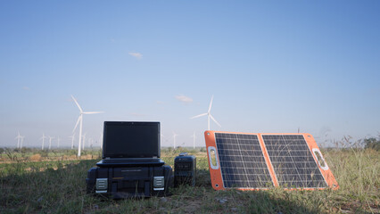 A foldable solar panel, laptop, and power station are set up in a grassy field surrounded by wind turbines, showcasing an off-grid renewable energy monitoring system under clear blue skies.