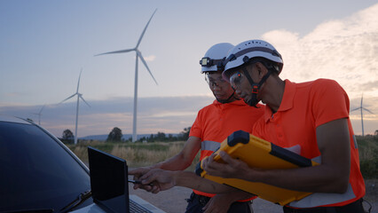 Two wind turbine engineers in safety gear analyze performance data using a laptop during sunset at a wind farm. Wind Energy Engineers Inspecting Turbines